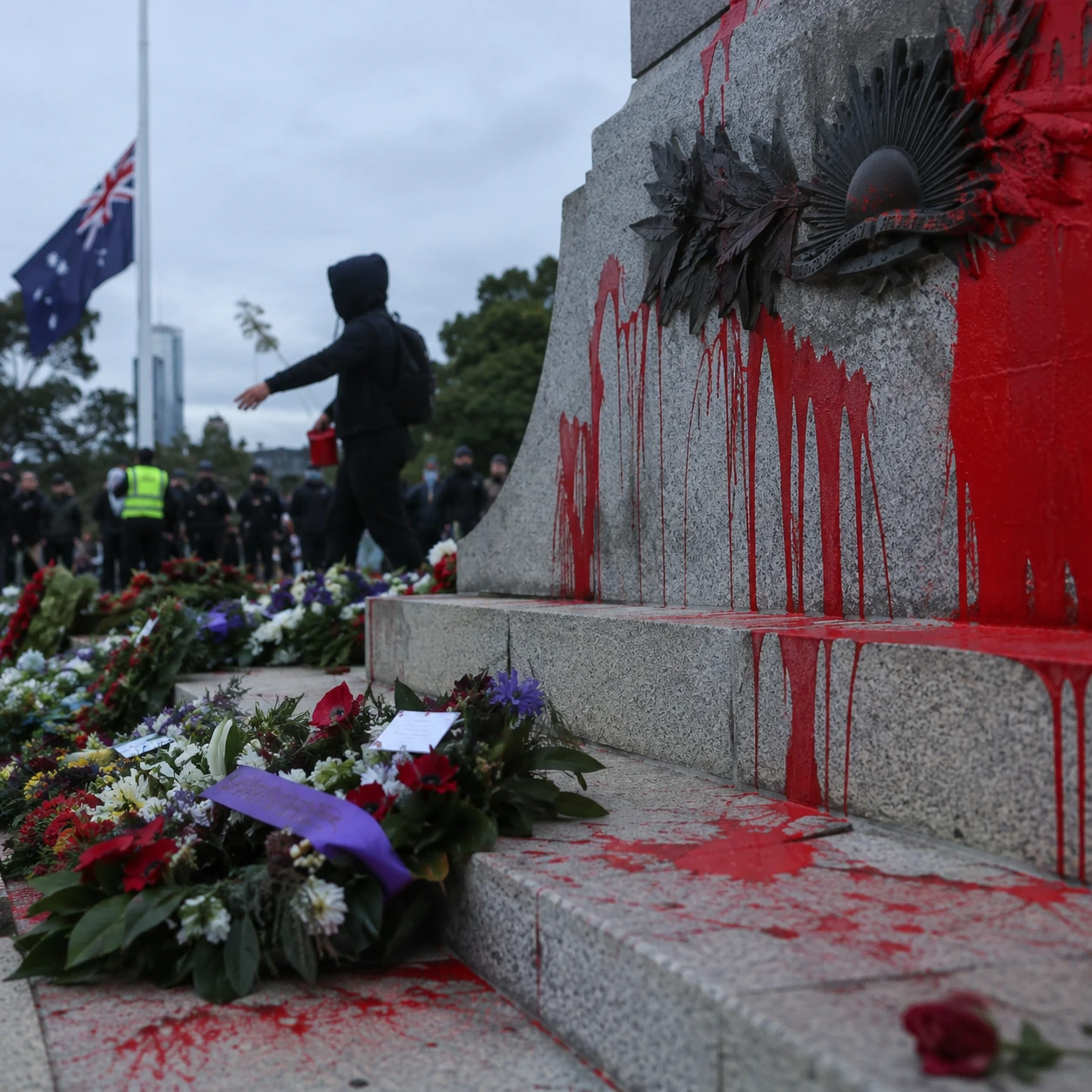 A war memorial covered in red paint with wreaths and a hooded figure nearby