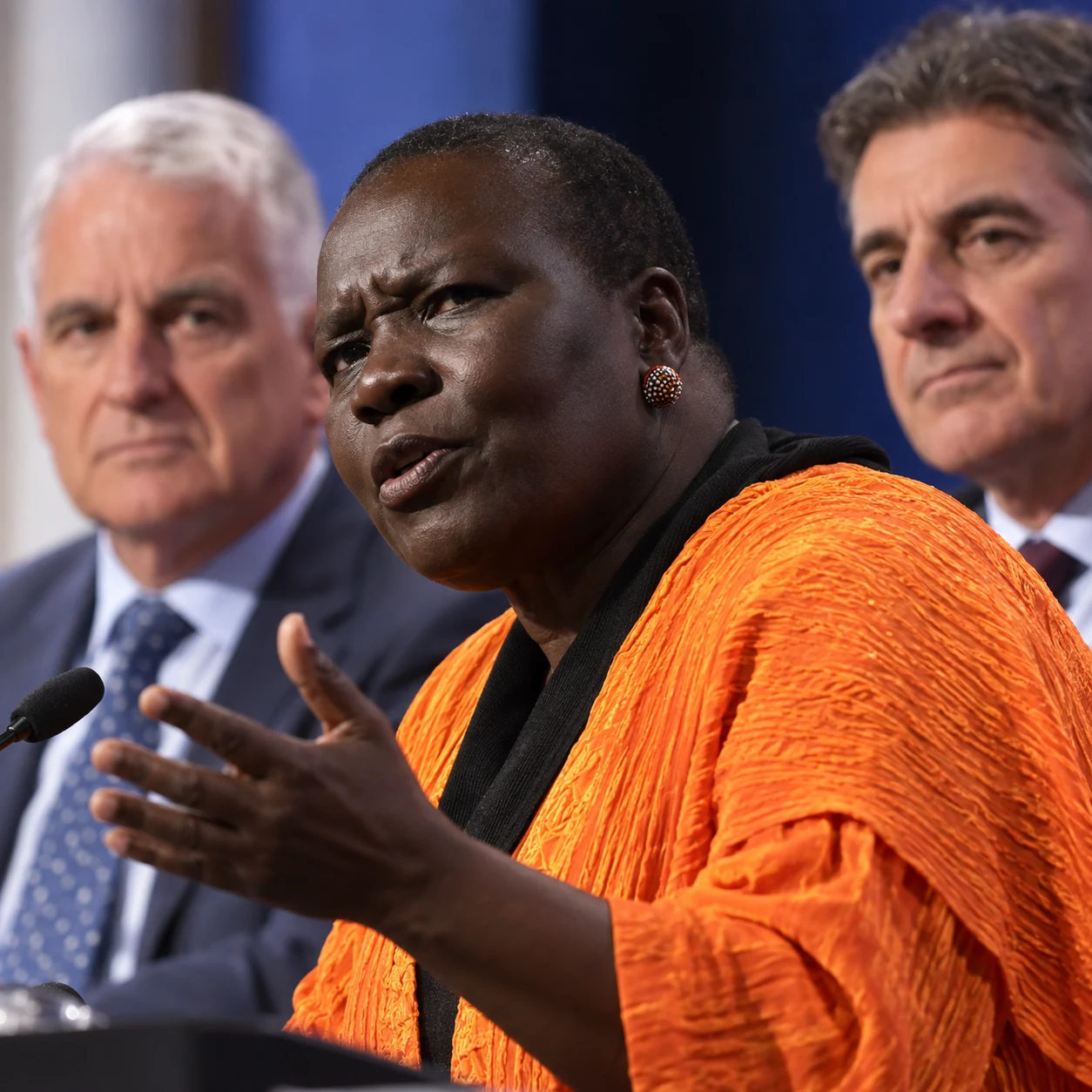 Woman speaking at an Australian public forum with two men seated behind her