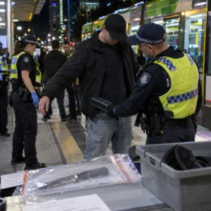 Police conducting a public safety search at a Melbourne transport stop at night.