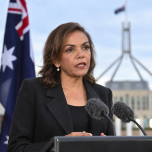 AI-generated conceptual illustration of a generic female politician at a lectern with Parliament House behind her; not a real photograph of Anne Aly or a documented press conference.
